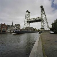 Overzicht, geklonken stalen brug, over de Gouwe, heeft twee taps toelopende heftorens met open vakwerkconstructie in Boskoop in 2003. Bron: Wikipedia.