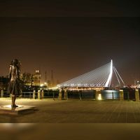 Rotterdam: Erasmusbrug bij avond, gezien vanaf de hoek Veerhaven-Westerkade. Het bronzen standbeeld van tsaar Peter de Grote gemaakt door Leonid Baranov in 1997 kijkt naar de brug.in 2008. Bron: Wikipedia.